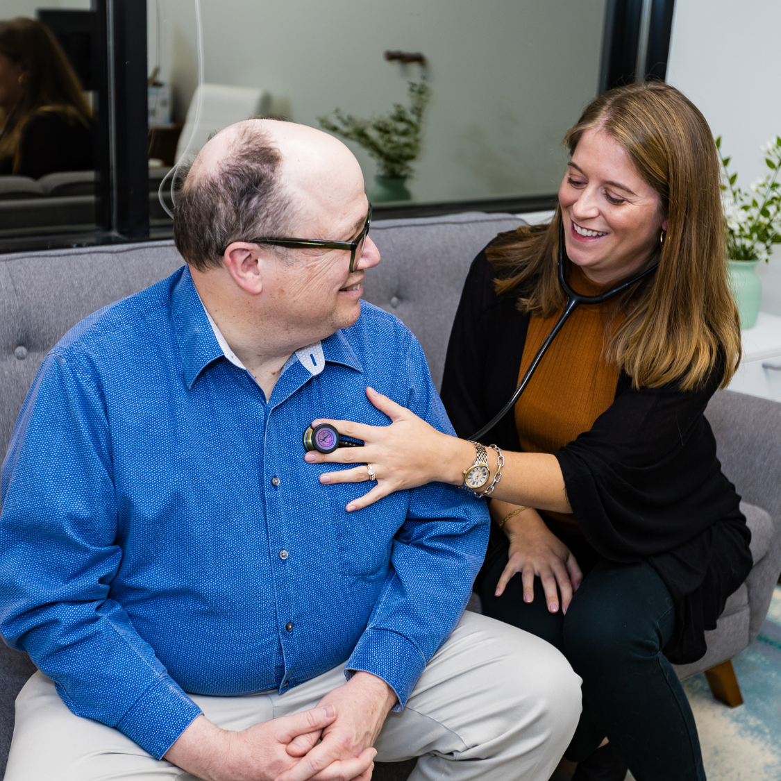 Female clinician with stethoscope listening to a smiling patient on a couch in a medical office.